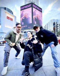 Three people pose energetically in front of a large outdoor advertisement on an urban street, with tall buildings in the background and cloudy skies above.